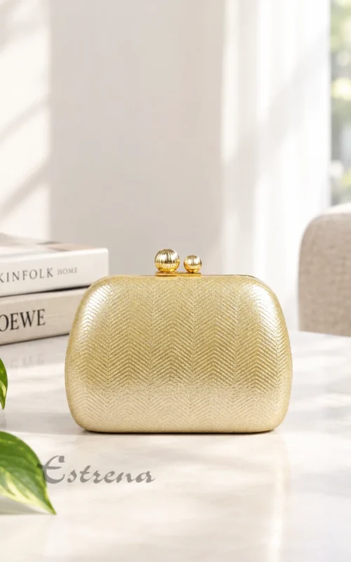 Bolso clutch rígido de fiesta color dorado con textura de espiga sobre una mesa junto a libros decorativos.