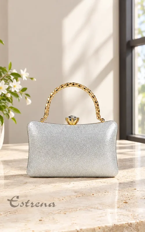 Bolso de fiesta plateado rígido con asa metálica dorada y cierre en forma de joya sobre una mesa.