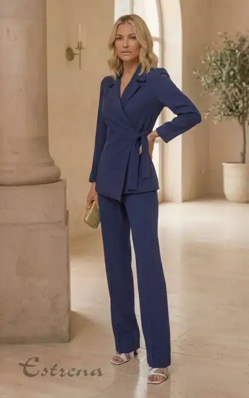 Mujer rubia con traje de chaqueta cruzada azul y lazo lateral, combinado con pantalón recto y bolso de mano dorado.