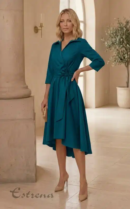 Mujer rubia con vestido midi camisero cruzado color azul verdoso con lazada y zapatos de tacón nude en un salón.