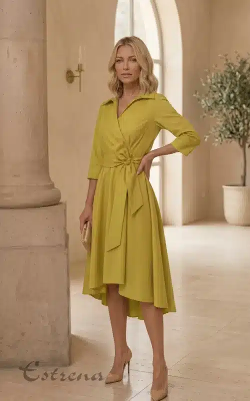Mujer rubia con vestido midi camisero cruzado color amarillo mostaza con lazo y zapatos de tacón nude en un gran salón.